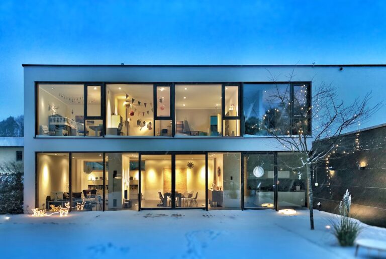 Maison contemporaine éclairée à la tombée de la nuit sous la neige, grandes baies vitrées et pièces chauffées